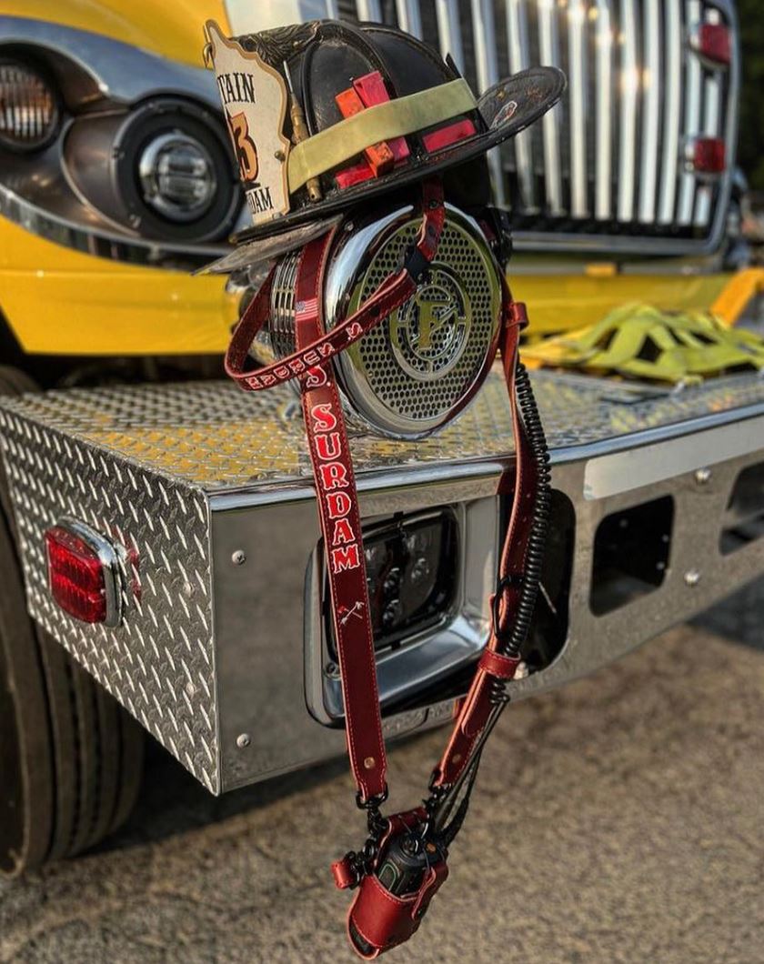 A custom JP radio strap draped over a fire helmet on the chrome bumper of a rig